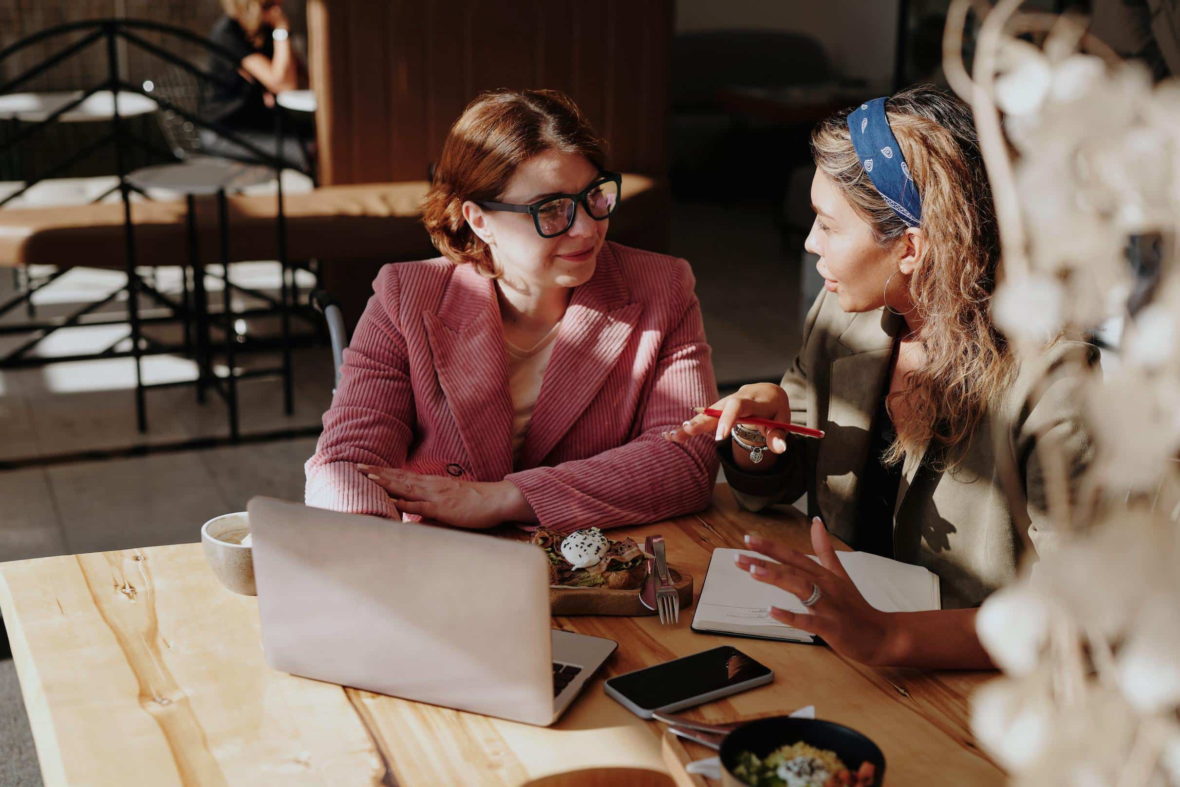Two women engaging in conversation at a coffee shop with a laptop and papers, practicing Portuguese in a casual and immersive setting.
