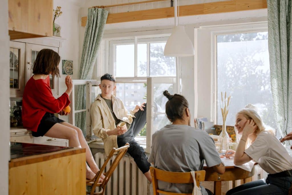 A group of young adults sitting around a dining table in a shared apartment, representing co-living, communication, and flatmate relationships.