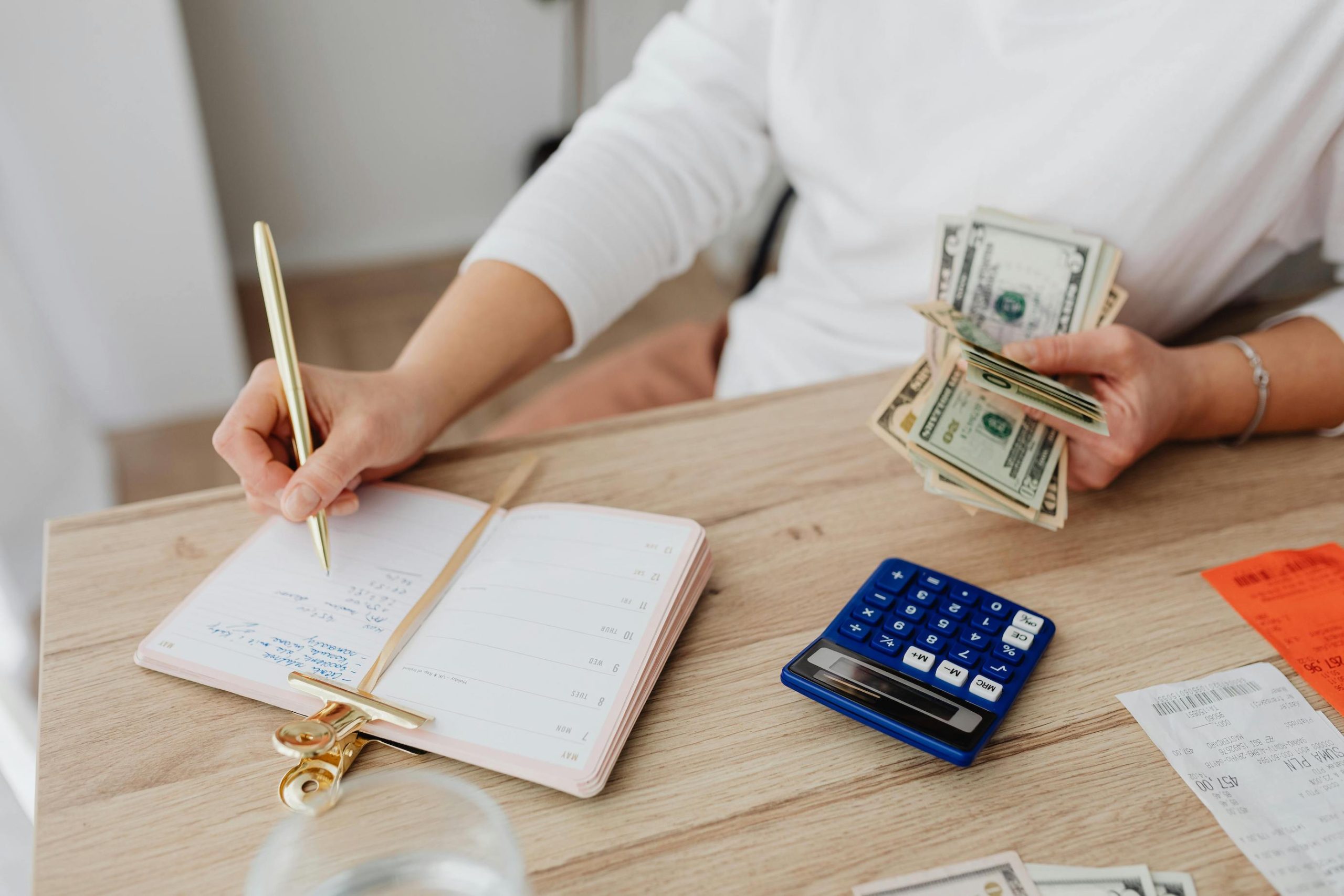 A person writing on a notebook while holding money, with a calculator on the side, symbolizing budgeting and cost-saving strategies for rent and utilities.