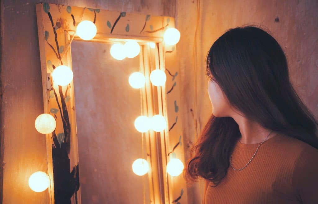 A woman gazing into a warmly lit mirror surrounded by decorative bulbs, showcasing DIY mood lighting ideas for a cozy home ambiance.