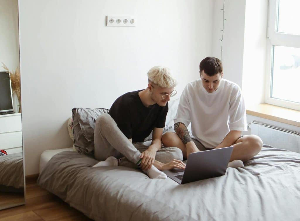 Two men sitting on a bed using their laptop, managing shared expenses and splitting bills in a shared apartment.