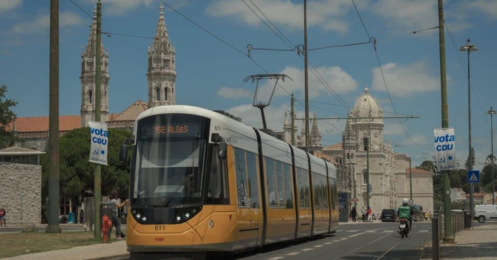 Yellow tram travels by Lisbon's iconic Jerónimos Monastery under clear blue skies, showcasing the city's historic public transport and cultural landmarks.