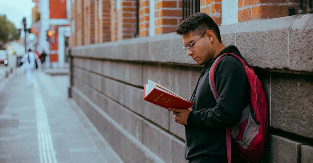 A young man leans against a brick wall while reading a red book on an urban street, symbolizing student life and education in Germany.