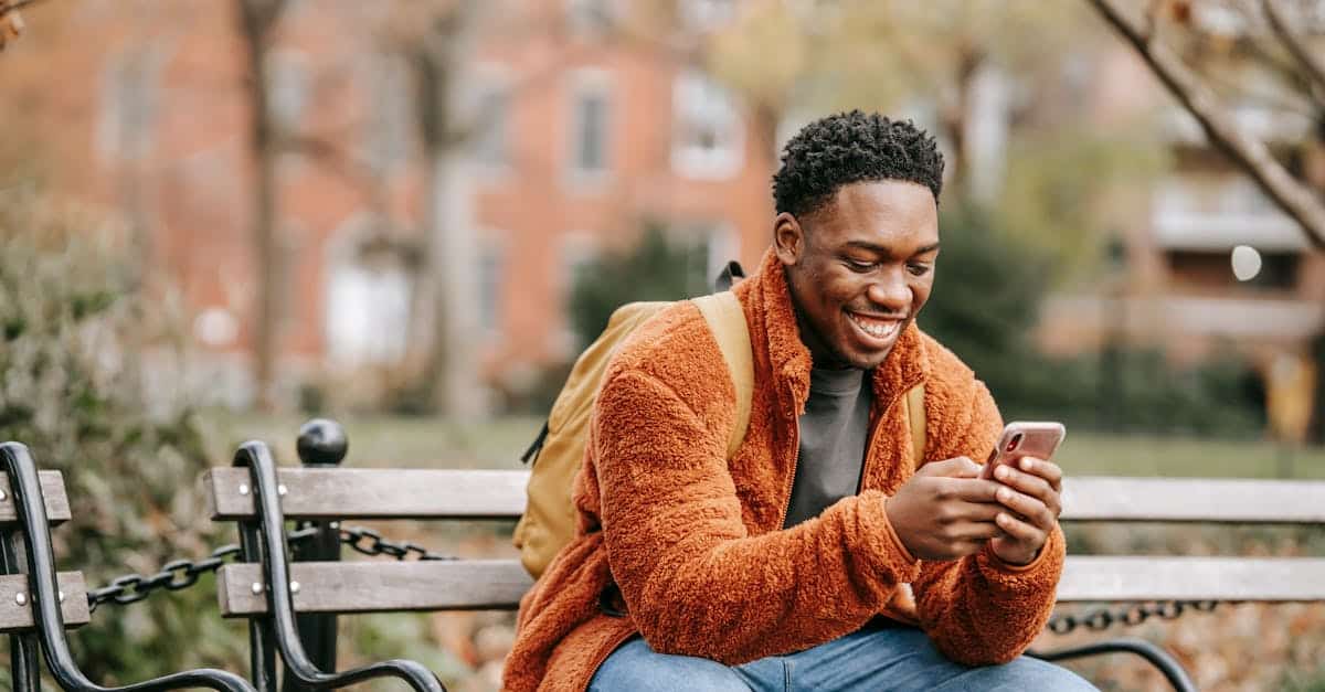 A cheerful young man in warm clothes smiling while using his phone on a bench in a city park, symbolizing easy navigation with Berlin transport apps.