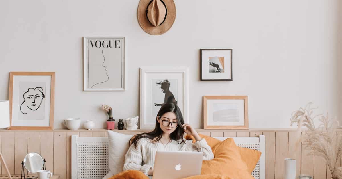 A young woman in a cozy bedroom using a laptop, surrounded by stylish decor and art, showcasing budget-friendly DIY decor ideas for small spaces.
