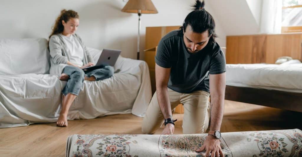A fit ethnic male in a wristwatch laying a carpet on parquet while a barefoot woman browses the internet on a portable computer sitting on a cozy sofa in a bedroom, illustrating budget-friendly rental furnishing ideas.