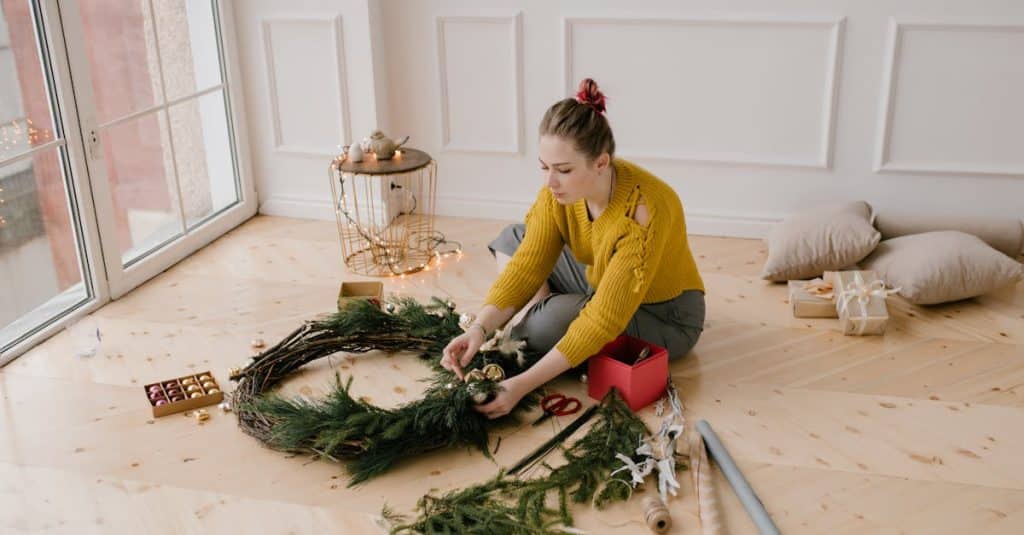 A young woman in a cozy room crafting a festive Christmas wreath by the window, showcasing budget-friendly DIY holiday decor ideas.