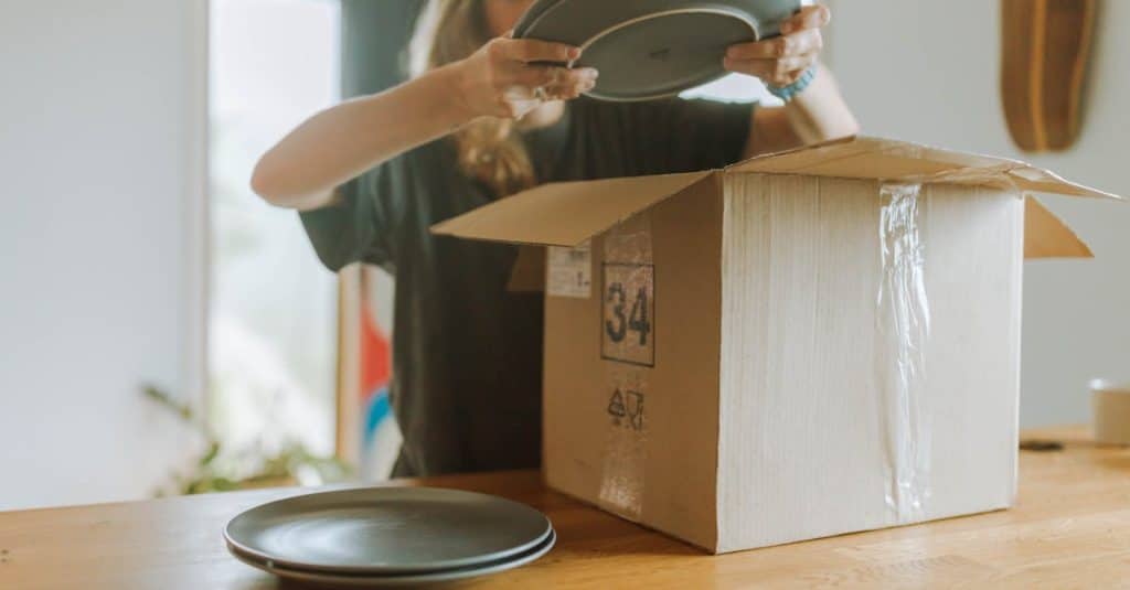 A woman packing ceramic plates into a cardboard box on a wooden table, preparing to move out of her shared apartment.