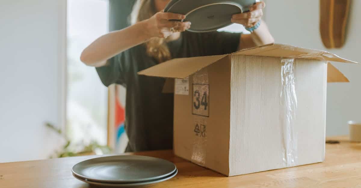 A woman packing ceramic plates into a cardboard box on a wooden table, preparing to move out of her shared apartment.