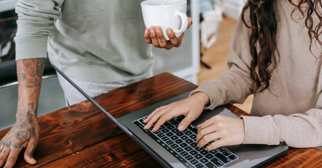 Two people working together at home, one typing on a laptop, the other holding coffee, symbolizing research and verification to avoid online rental scams.