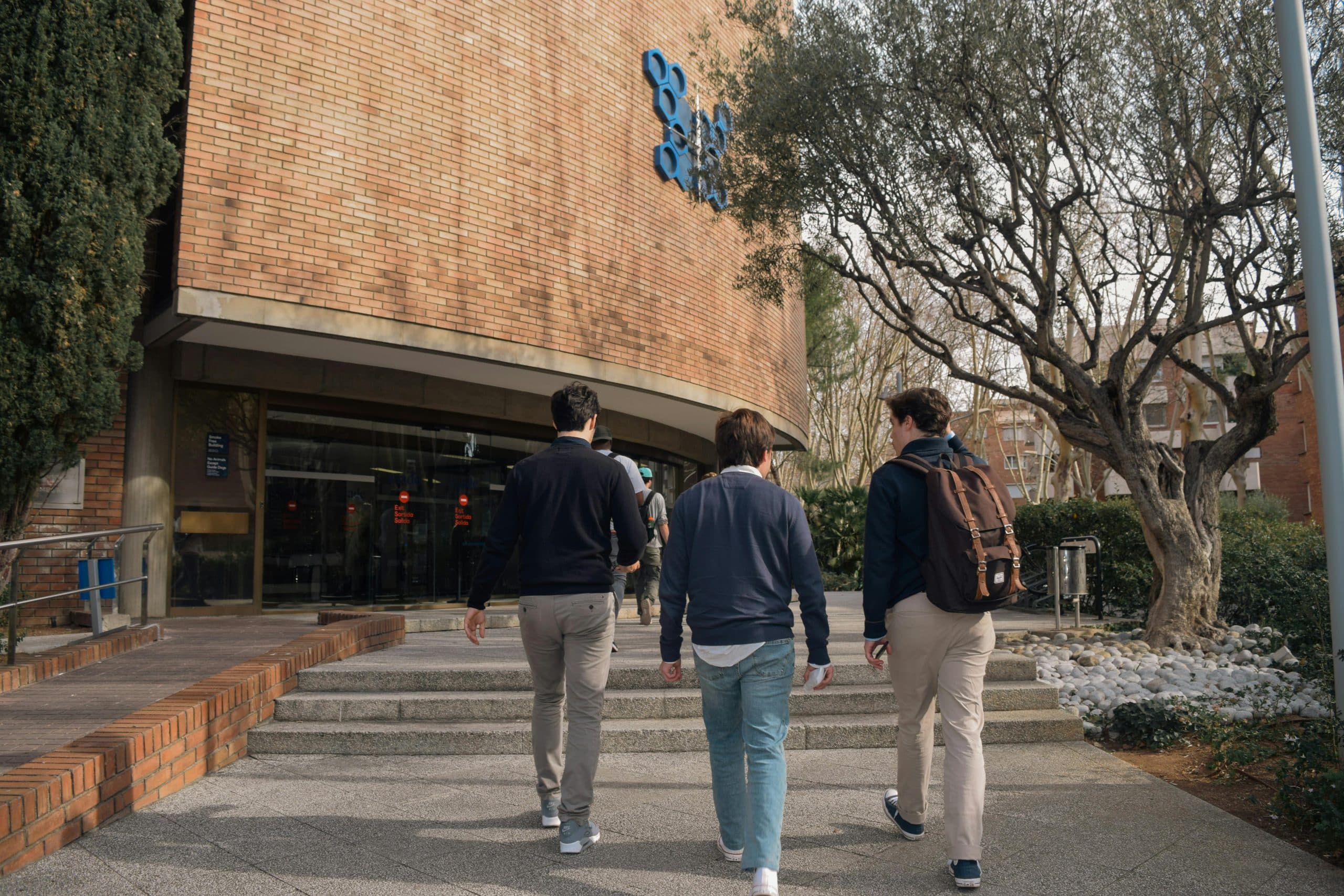 Three students approach a university building entrance in Barcelona, Spain on a sunny day.
